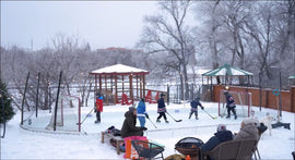Multiple kids playing on a backyard hockey rink while spectators watch by a fire pit on a snowy day.