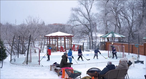 Multiple kids playing on a backyard hockey rink while spectators watch by a fire pit on a snowy day.