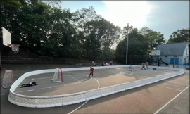 Children play street hockey inside a white portable rink enclosure set up on an outdoor concrete basketball court.
