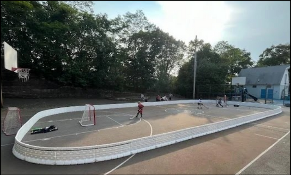 Children play street hockey inside a white portable rink enclosure set up on an outdoor concrete basketball court.