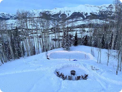 A backyard ice rink on a very snowy day next to five chairs and a fire pit