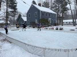 Three kids playing ice hockey on a YardRink backyard ice rink with three people watching from chairs on the outside of it.