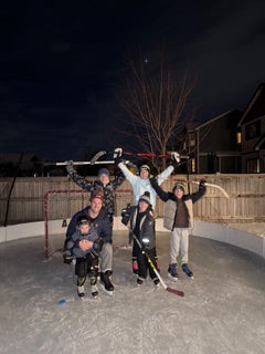A family poses together with raised hockey sticks on an illuminated backyard ice rink at night.