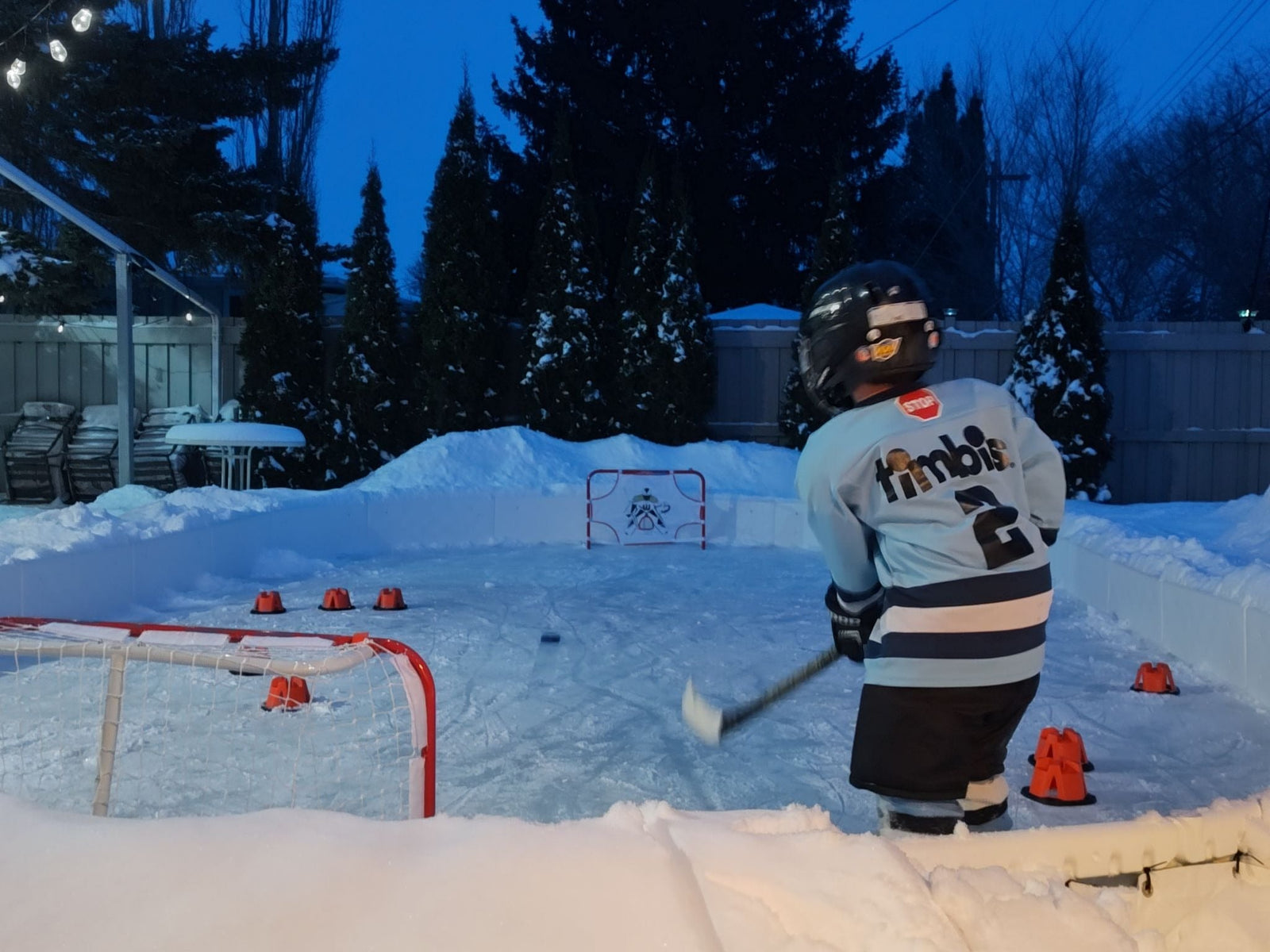 A young child wearing a hockey helmet and a light blue "Timbits" jersey shoots a puck on a snow-bordered backyard ice rink.
