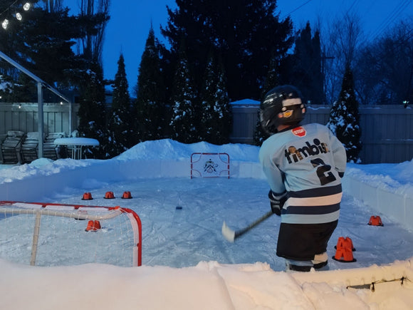 A young child wearing a hockey helmet and a light blue "Timbits" jersey shoots a puck on a snow-bordered backyard ice rink.