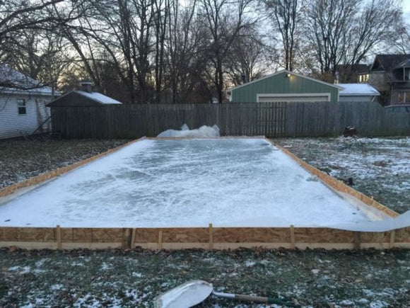 A rectangular homemade backyard ice rink under construction, featuring a frozen ice surface contained by wooden boards and a plastic liner in a grassy yard.