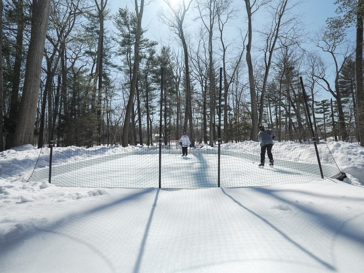 Two kids skate and play hockey behind a protective mesh net on a snow-surrounded backyard ice rink in.