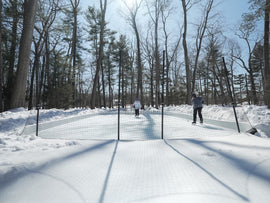 Two kids skate and play hockey behind a protective mesh net on a snow-surrounded backyard ice rink in.