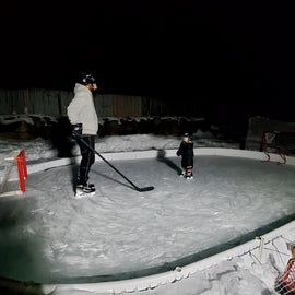 A man and little boy standing across from each other on a backyard ice rink at night.