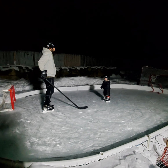 A man and little boy standing across from each other on a backyard ice rink at night.