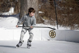 A young boy holding a hockey stick on a backyard ice rink with a Boston Bruins logo on a panel.