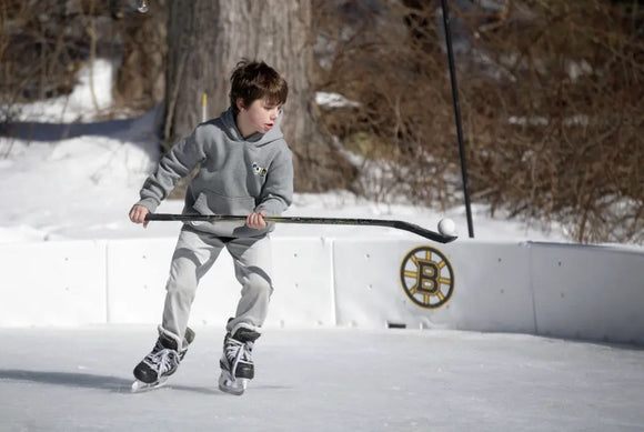 A young boy holding a hockey stick on a backyard ice rink with a Boston Bruins logo on a panel.