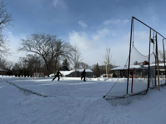 Two people skate and play ice hockey on a large, snowy backyard ice rink equipped with a tall netting system.