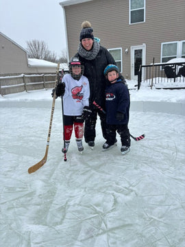 An adult and two young children wearing helmets and ice skates smile together while standing on a textured backyard ice rink.