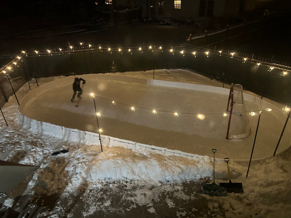  A backyard ice rink illuminated by string lights at night, featuring a person playing hockey and a red goal net surrounded by snow.