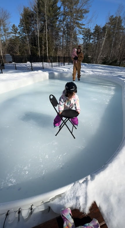 A young child wearing a helmet and winter gear balances while holding a folding chair on a backyard ice rink, with an adult holding a toddler ice skating in the background