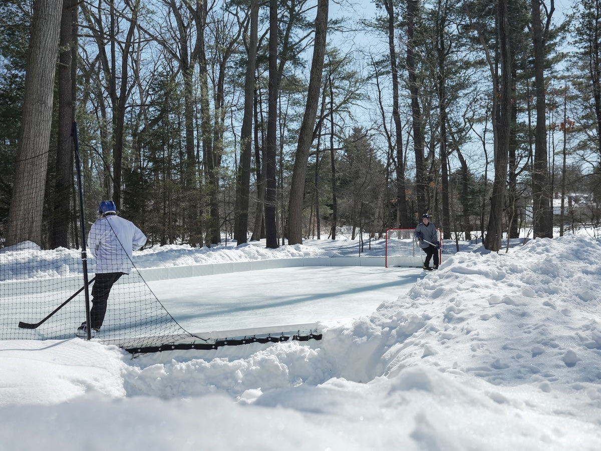 Two kids playing hockey on a YardRink backyard ice rink with piles of snow outside of the rink.