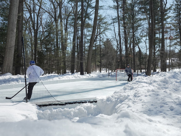 Two kids playing hockey on a YardRink backyard ice rink with piles of snow outside of the rink.