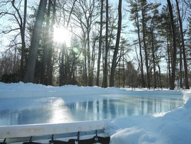 Sunlight gleams off the smooth ice surface of a backyard skating rink surrounded by snowy banks and tall trees.