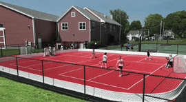 Multiple kids playing street hockey on a red court next to two buildings.