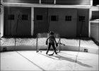 A black and white photo of a youth hockey player standing in front of a hockey net on a YardRink home ice rink system, featuring an NHL logo badge on the rink boards and a tall backstop safety net.