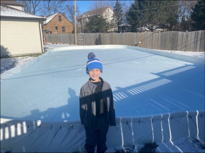 A smiling young boy wearing a winter hat standing in front of a large, frozen YardRink backyard ice rink set up in a suburban residential backyard with a wooden fence and houses in the background.