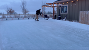 An adult practicing hockey stick handling on a large YardRink home ice rink system set up next to a patio with string lights, featuring official NHL team logo decals on the white rink boards.